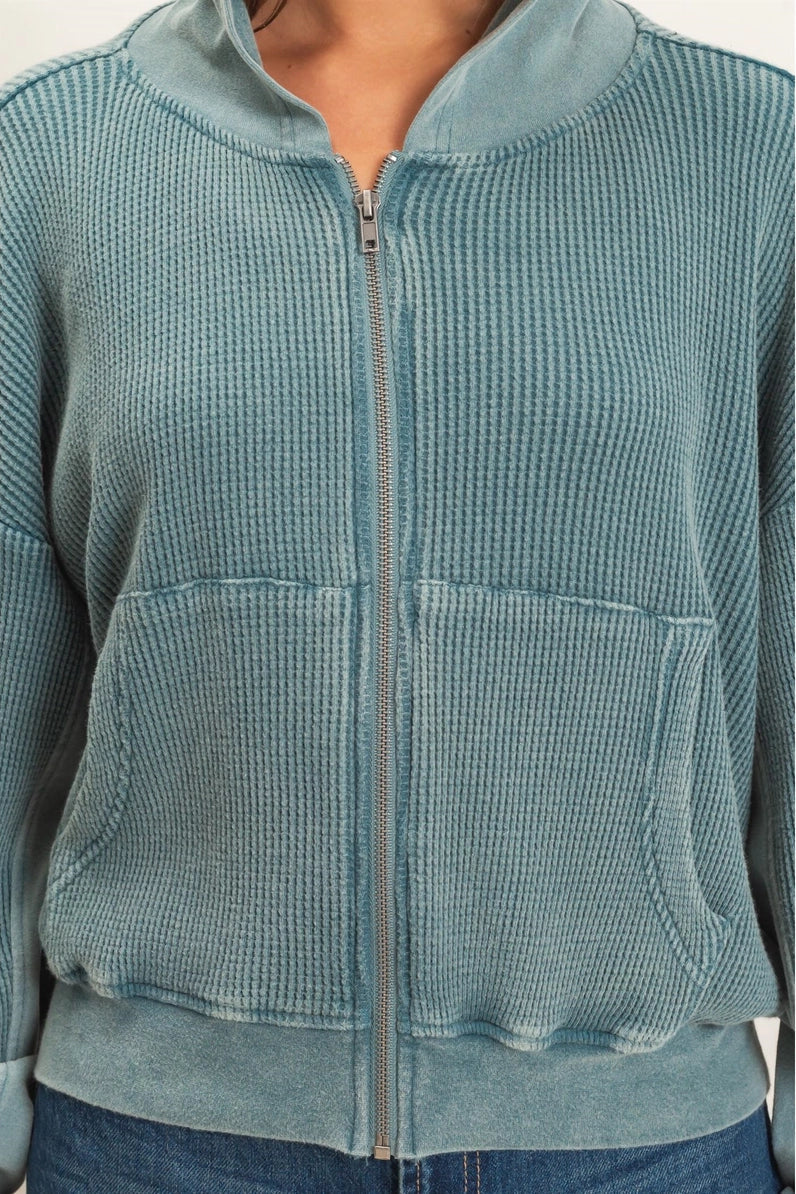 A cropped, ribbed-texture zip-up jacket in a dusty teal blue-green color, featuring a stand collar, front zipper, and two large front patch pockets, shown on a model.