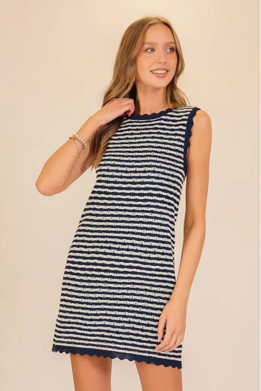 A woman modeling a sleeveless knit mini dress with horizontal navy and off-white stripes, featuring a scalloped neckline and hem, set against a plain beige background.