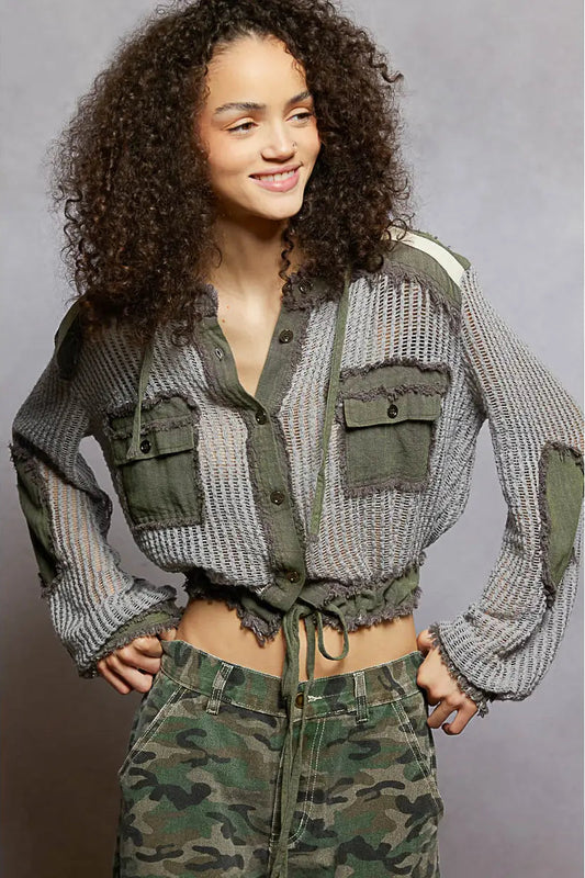 A woman modeling a cropped, long-sleeve button-front shirt with a textured gray and olive green knit pattern, frayed edges, and flap chest pockets, paired with camouflage pants against a neutral gray studio background.