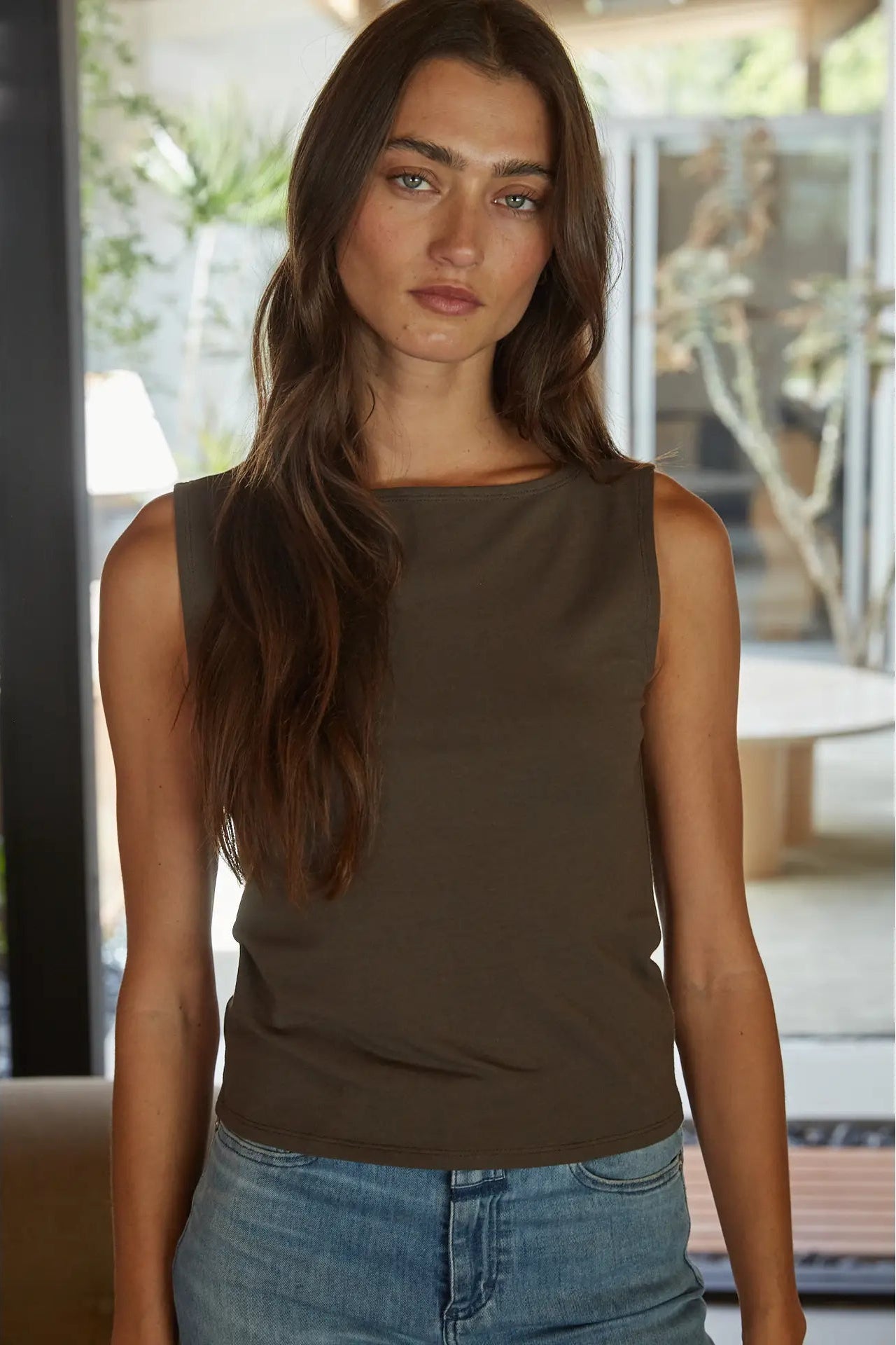 A woman wearing a sleeveless, boat-neck tank top in a solid dark brown color, paired with light-wash blue jeans, standing in a modern indoor setting with a blurred background.