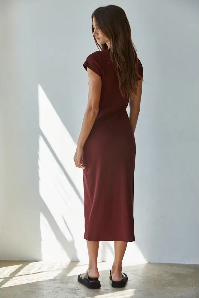 A woman modeling a short-sleeve, knee-length burgundy knit dress with black slide sandals, standing in a brightly lit room with diagonal sunlight shadows on a white wall.