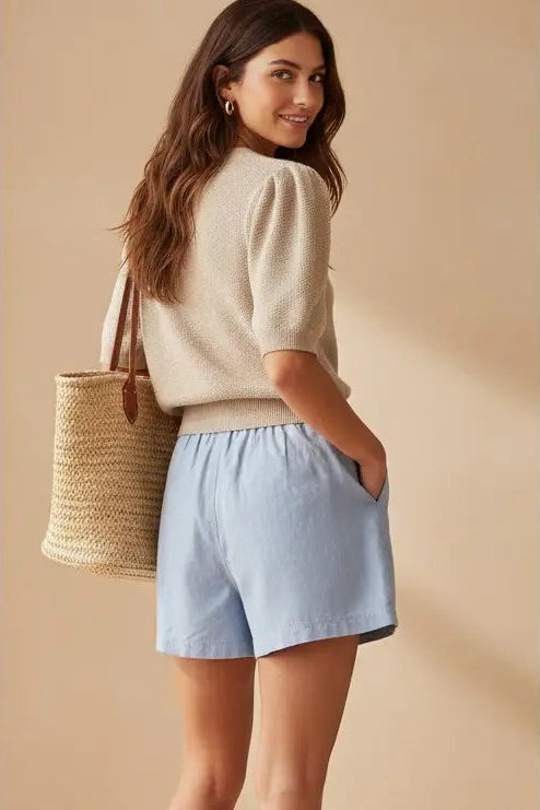 Woman wearing a beige sweater and light blue shorts, holding a straw bag against a beige background