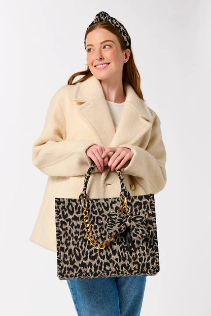 A woman wearing a cream coat, blue jeans, and a leopard-print headband holds a leopard-print tote bag with gold chain handles against a light gray studio background.