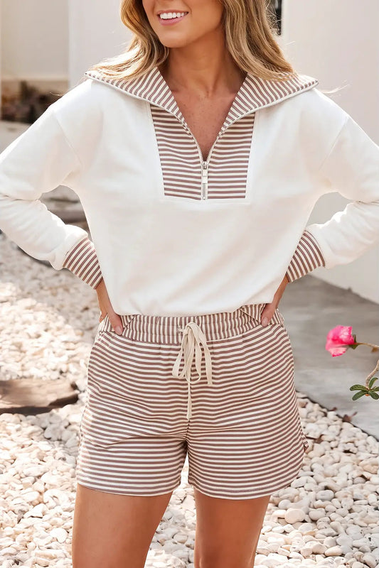 A woman wearing a matching two-piece lounge set with a white long-sleeve half-zip top and beige-and-white horizontally striped shorts, standing outdoors on a pebble surface.