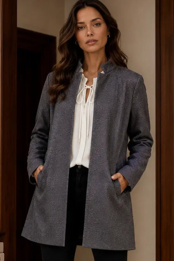 A woman wearing a mid-length gray textured coat over a white tie-front blouse and black pants, standing indoors in front of a dark wood door frame.