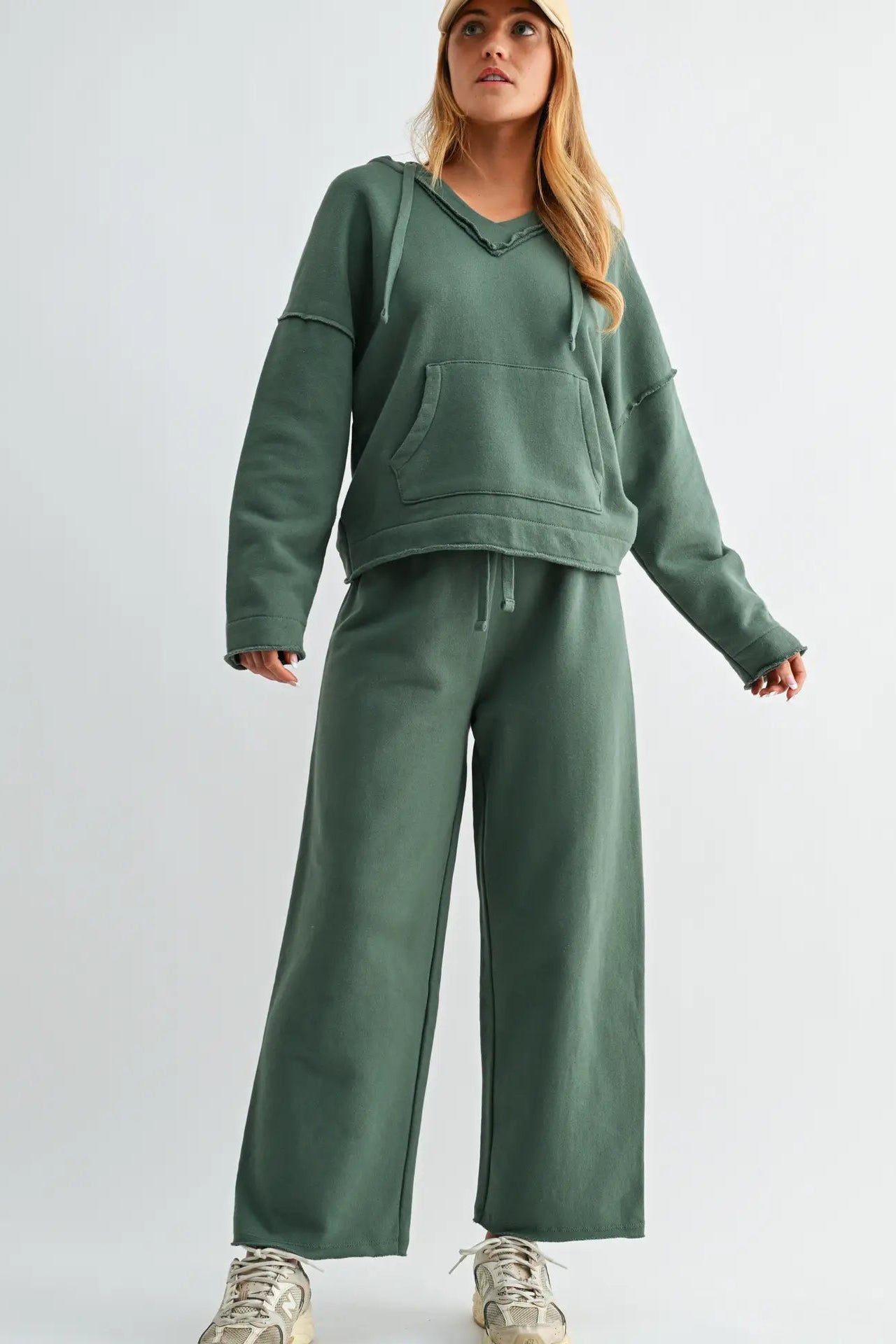 A woman modeling a matching sage green lounge set consisting of a cropped pullover hoodie with a kangaroo pocket and wide-leg sweatpants, paired with beige sneakers, against a light gray studio background.