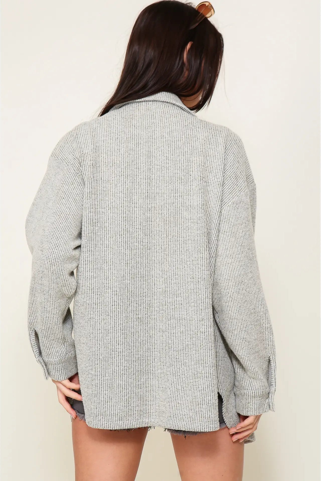 A woman viewed from behind wearing an oversized light gray ribbed knit button-up shirt jacket with raw, frayed edges.