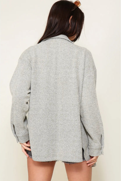 A woman viewed from behind wearing an oversized light gray ribbed knit button-up shirt jacket with raw, frayed edges.