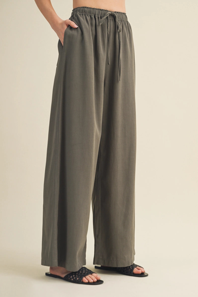 A person wearing loose, wide-leg drawstring pants in a muted olive-gray color, paired with black slide sandals, against a plain beige studio background.