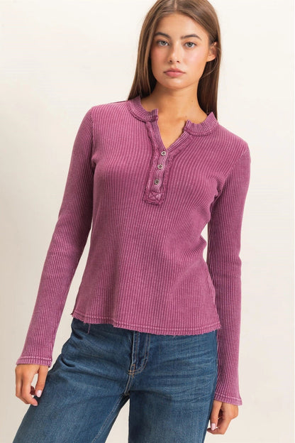 A woman modeling a ribbed long-sleeve henley top in a dusty mauve-purple color, paired with blue jeans, against a plain light beige background.
