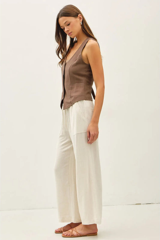A woman modeling a sleeveless brown vest paired with wide-leg cream pants and flat sandals, standing against a plain off-white studio background.
