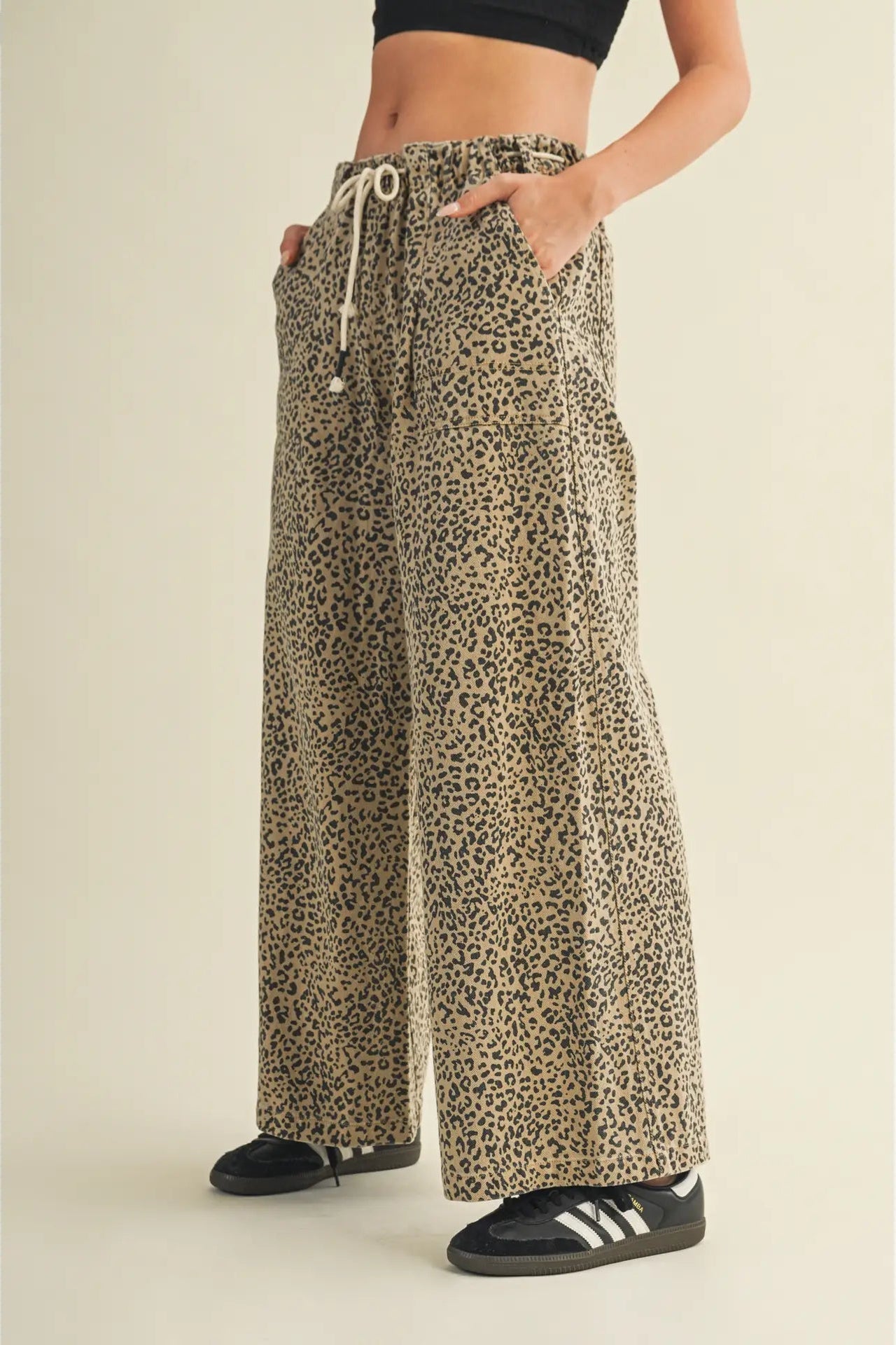 A person wearing loose, wide-leg leopard-print pants with a drawstring waist, paired with black sneakers, shown against a plain beige studio background.