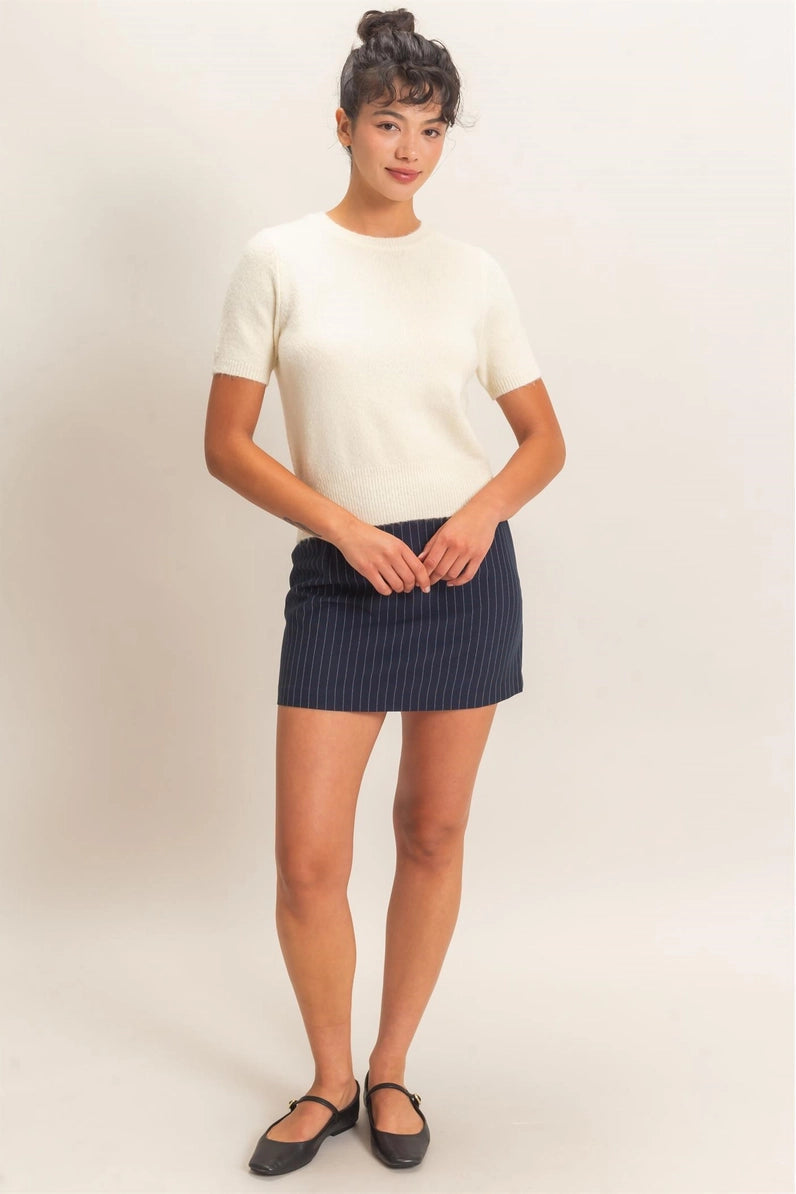A woman modeling a cream short-sleeve knit top and a dark navy blue pleated mini skirt, paired with black flats, against a plain light beige studio background.