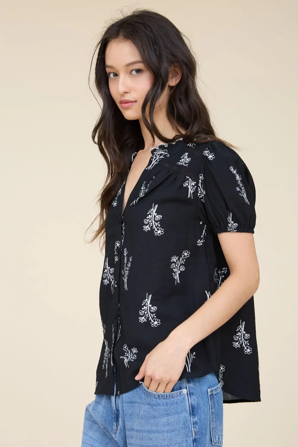 A woman wearing a black short-sleeve button-up blouse with a white floral print, paired with blue jeans, photographed against a solid beige studio background.