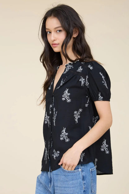 A woman wearing a black short-sleeve button-up blouse with a white floral print, paired with blue jeans, photographed against a solid beige studio background.