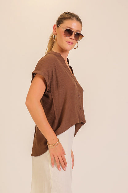 A woman wearing a brown short-sleeve top and white pants, photographed against a plain light beige studio background.