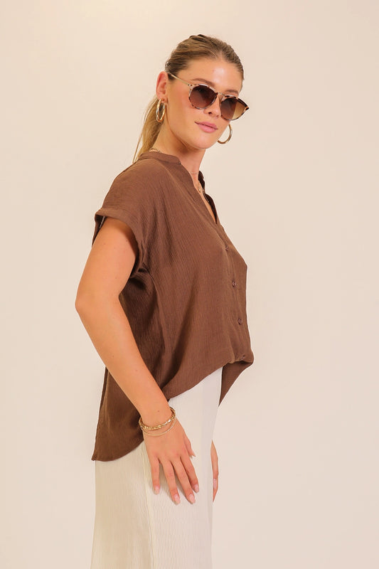 A woman wearing a brown short-sleeve top and white pants, photographed against a plain light beige studio background.