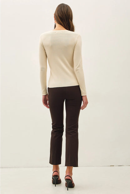 A woman viewed from behind wearing a cream long-sleeve knit top, dark brown cropped pants, and black heels against a plain white studio background.