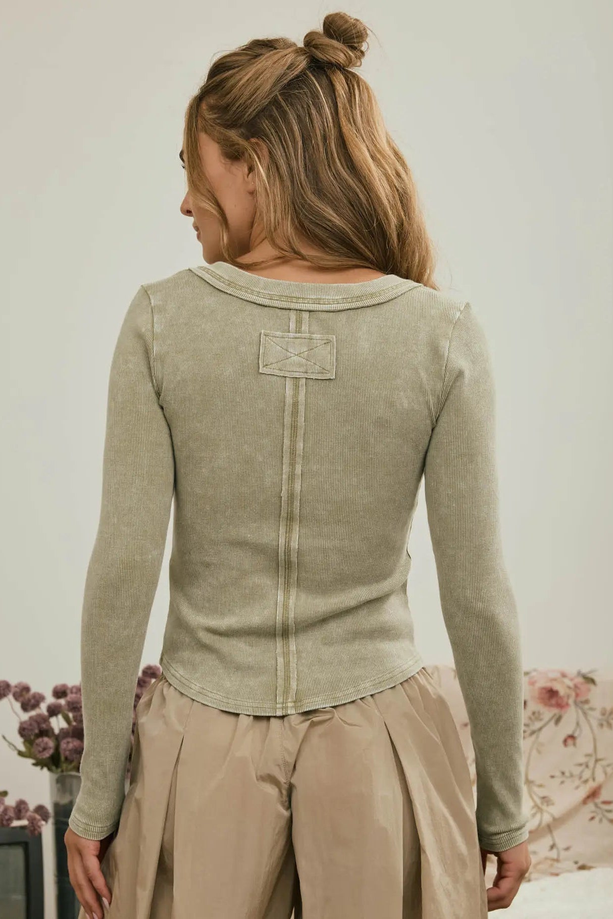 A woman shown from the back wearing a light sage green long-sleeve knit top with a decorative stitched patch on the upper back, paired with beige pleated pants in a softly lit indoor setting.