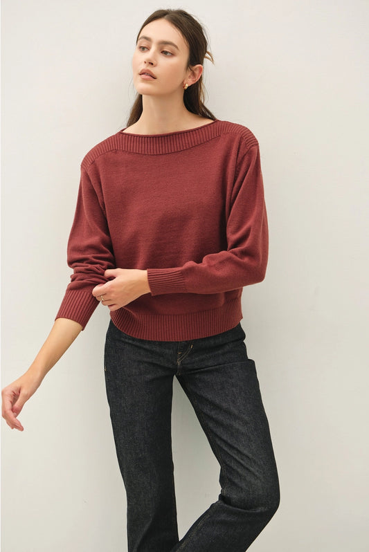 A woman modeling a rust-red knit sweater with a wide boat neckline and dark wash jeans, standing against a plain light gray wall.