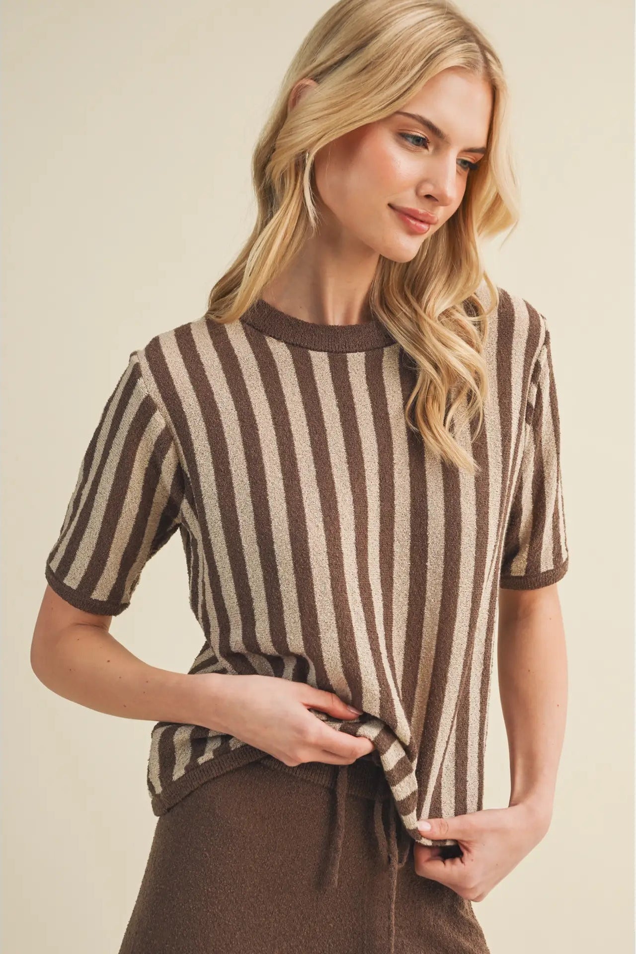 A woman wearing a brown and beige vertically striped short-sleeve knit top, styled with a matching brown skirt against a solid beige background.