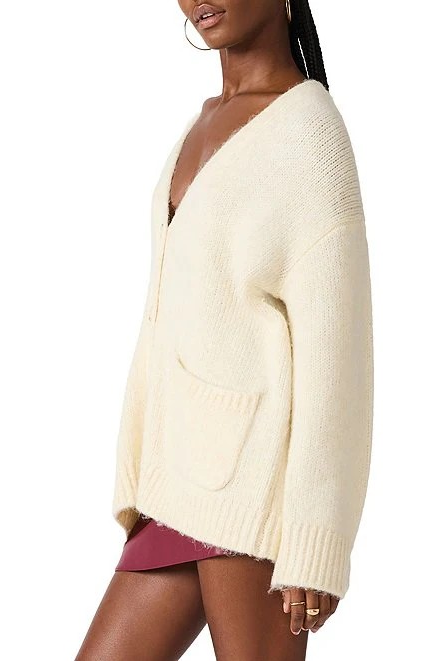 A woman wearing an oversized cream-colored knit cardigan with a deep V-neck, front pockets, and ribbed cuffs and hem, shown against a plain white background.