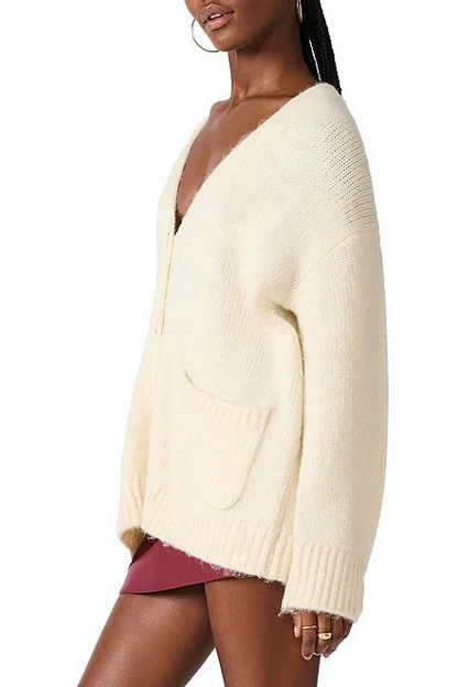 A woman wearing an oversized cream-colored knit cardigan with a deep V-neck, front pockets, and ribbed cuffs and hem, shown against a plain white background.