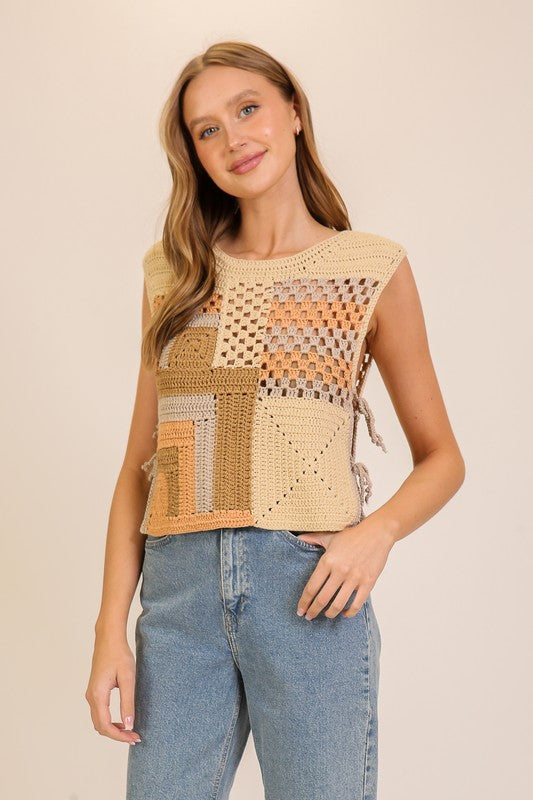 A woman modeling a sleeveless crochet knit top with a patchwork design in beige, tan, and brown tones, paired with light blue jeans against a solid light beige background.