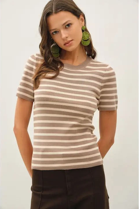 A woman modeling a short-sleeve ribbed knit top with horizontal taupe and beige stripes, paired with dark brown bottoms and large green earrings, against a plain light gray background.