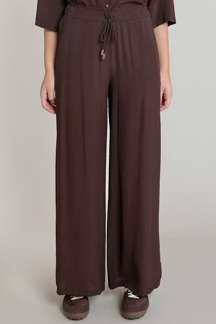 A person wearing dark brown wide-leg drawstring pants with a matching top and brown shoes, standing against a plain light gray studio background.