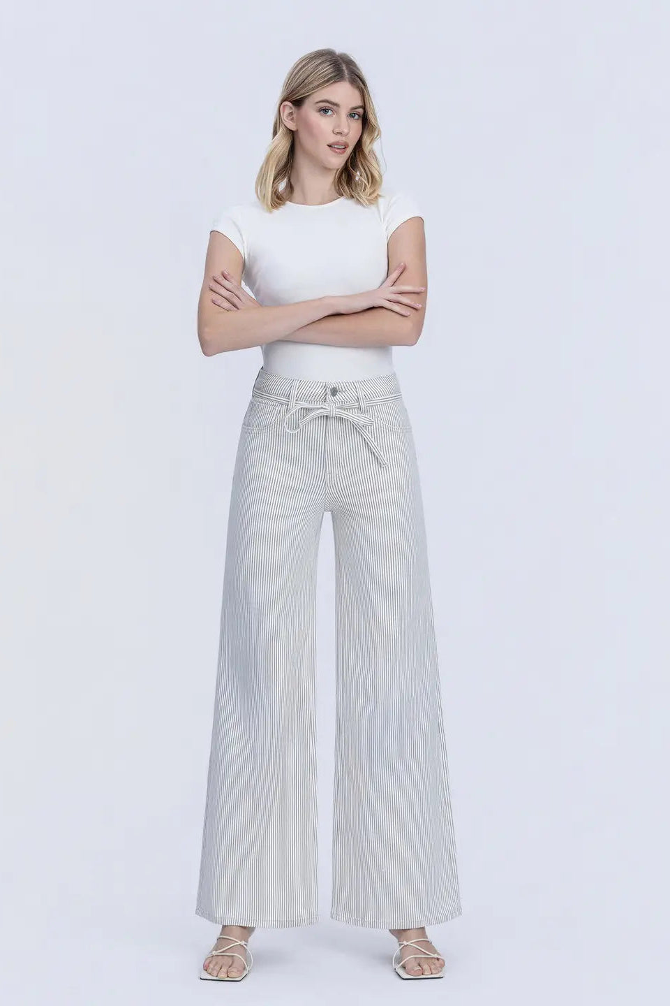A woman modeling a pair of light gray wide-leg pants with a drawstring waist, paired with a white short-sleeve top, against a plain light gray studio background.