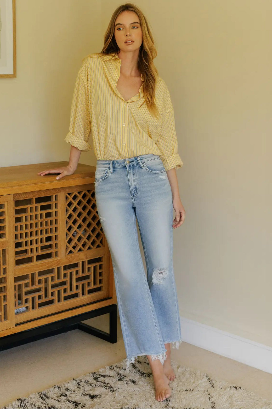 Woman in a yellow blouse and light blue jeans standing next to a wooden cabinet in a room.