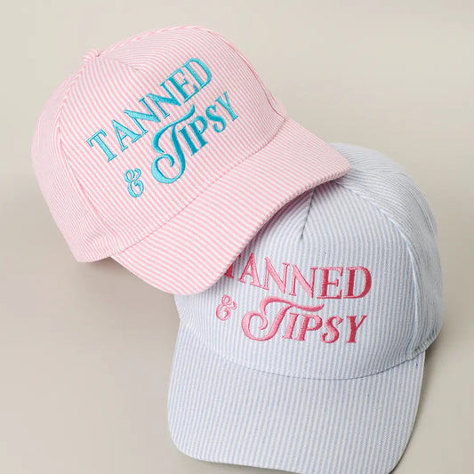 Two baseball caps with 'Tanned & Tipsy' text on a beige background
