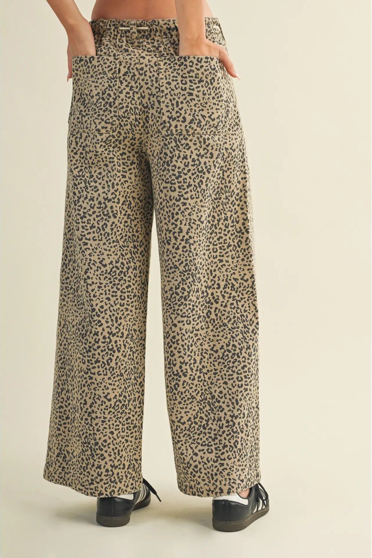 Wide-leg trousers with an all-over leopard print pattern in beige, brown, and black, shown on a model from the waist down wearing black shoes against a solid light beige background.