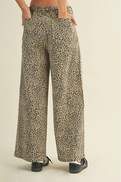 Wide-leg trousers with an all-over leopard print pattern in beige, brown, and black, shown on a model from the waist down wearing black shoes against a solid light beige background.