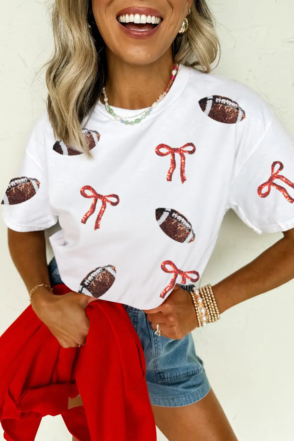 A woman wearing a white short-sleeve t-shirt with a pattern of red bows and brown footballs, paired with denim shorts and holding a red sweater.