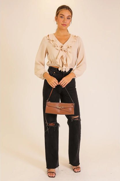 A woman modeling a beige ruffled long-sleeve blouse paired with black distressed jeans, brown sandals, and a brown shoulder bag against a plain beige studio background.