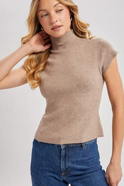 A woman wearing a fitted short-sleeve mock neck top in a heathered taupe-brown color, paired with blue jeans, against a light gray studio background.