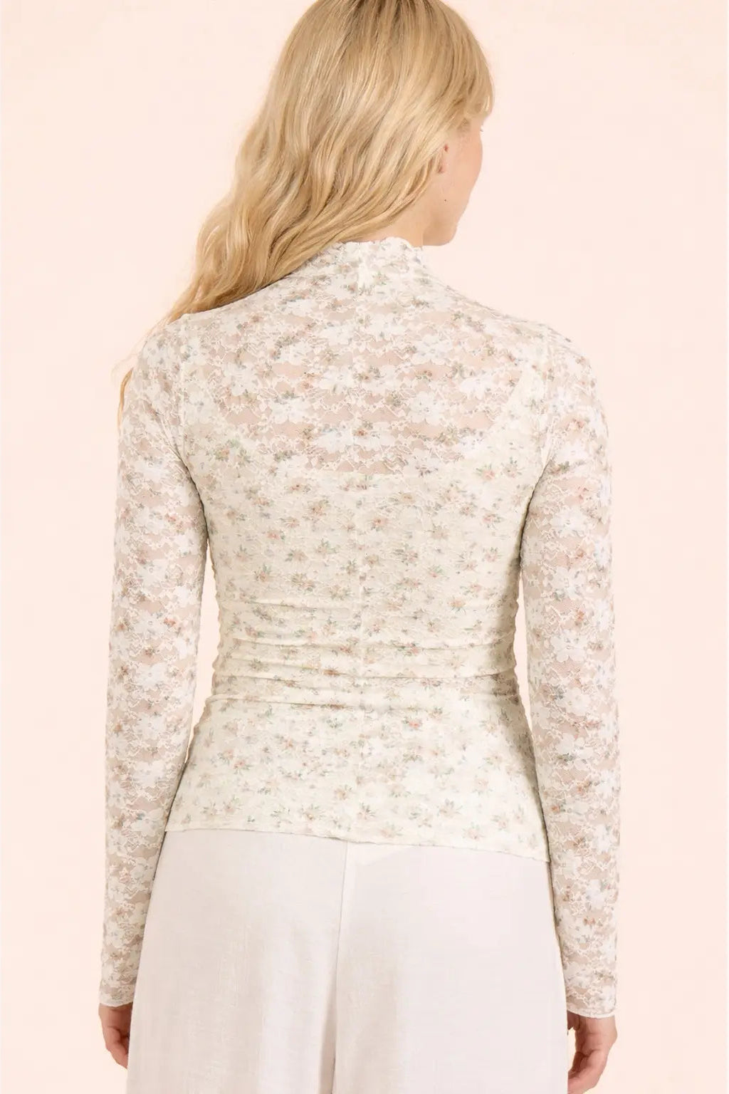 A woman shown from the back wearing a fitted long-sleeve mock-neck top with a delicate beige and white floral lace pattern, paired with light-colored pants, against a solid pale pink background.