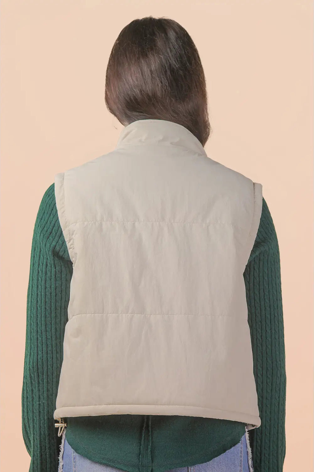 A person shown from the back wearing a light beige quilted puffer vest over a dark green ribbed long-sleeve top, against a solid pale peach background.