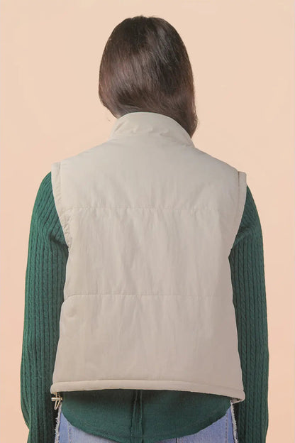 A person shown from the back wearing a light beige quilted puffer vest over a dark green ribbed long-sleeve top, against a solid pale peach background.