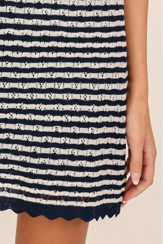 Close-up of a ribbed knit dress with horizontal navy and cream stripes, featuring a scalloped hem and a model's hand holding the side.