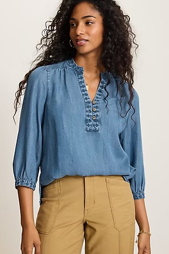 A woman wearing a light blue denim-like blouse with a buttoned split neckline and gathered sleeves, paired with tan pants, against a plain light beige studio background.