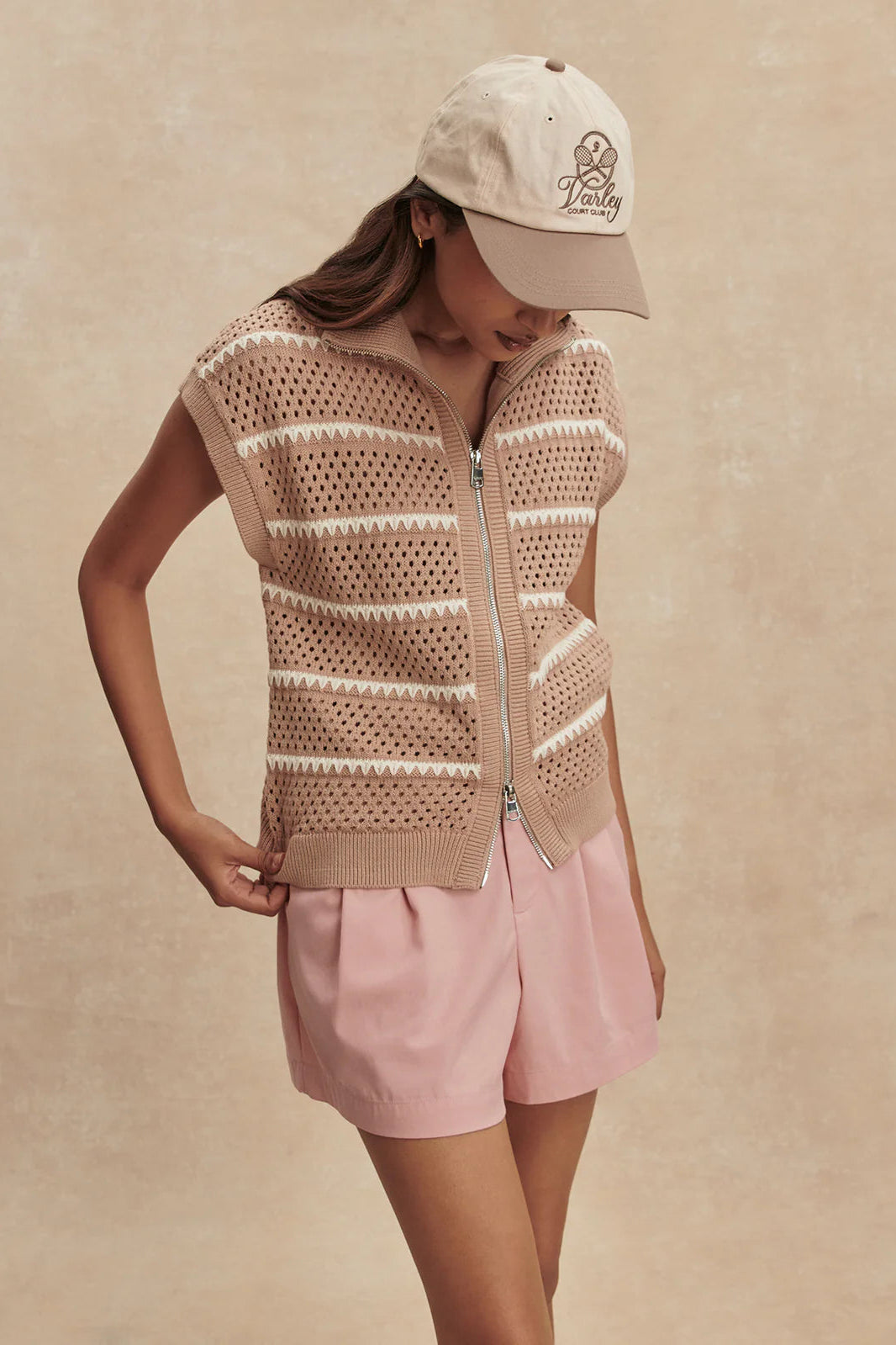 Person wearing a beige knitted vest and pink shorts with a beige cap on a beige background