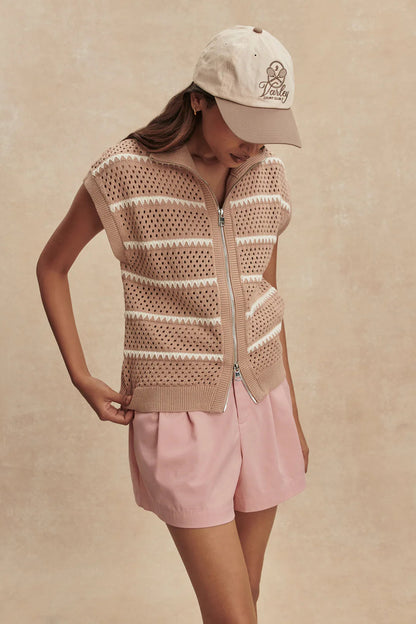 Person wearing a beige knitted vest and pink shorts with a beige cap on a beige background