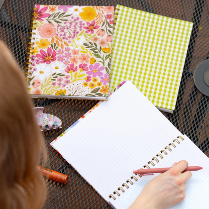 Person writing in a spiral-bound notebook with floral and checkered covers on a textured surface.