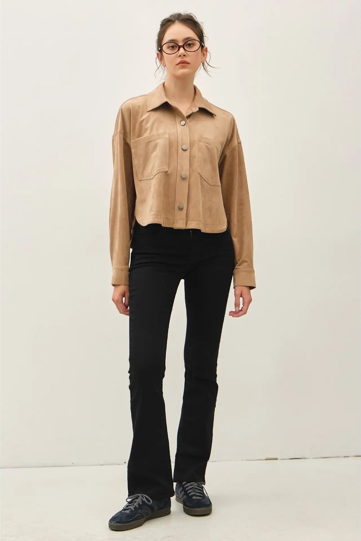 A model wearing a cropped tan suede-like button-up shirt with chest pockets, black flared jeans, and dark blue sneakers, standing against a plain off-white studio background.