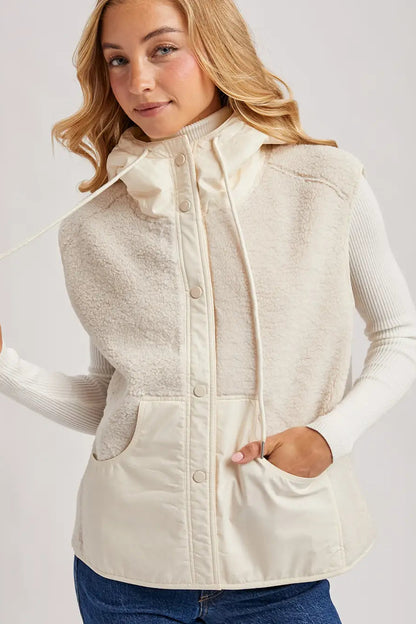 A woman wearing a cream-colored hooded fleece vest over a ribbed long-sleeve top and blue jeans, shown against a light gray studio background.