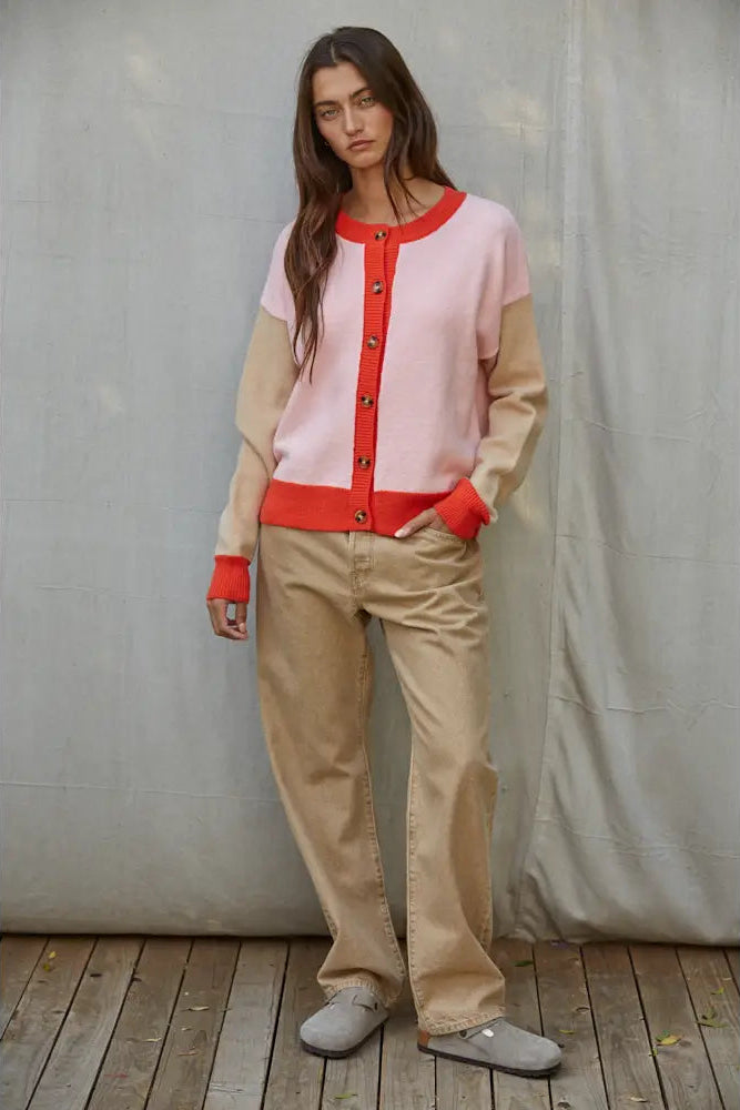 A woman stands against a draped gray fabric backdrop on a wooden floor, wearing a color-block cardigan with a light pink body, beige sleeves and hem, and a red button placket and cuffs, paired with beige pants and gray slip-on shoes.