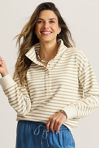 A woman wearing a cream and tan striped long-sleeve henley top with a mock neck and button placket, paired with blue drawstring pants, against a light gray studio background.
